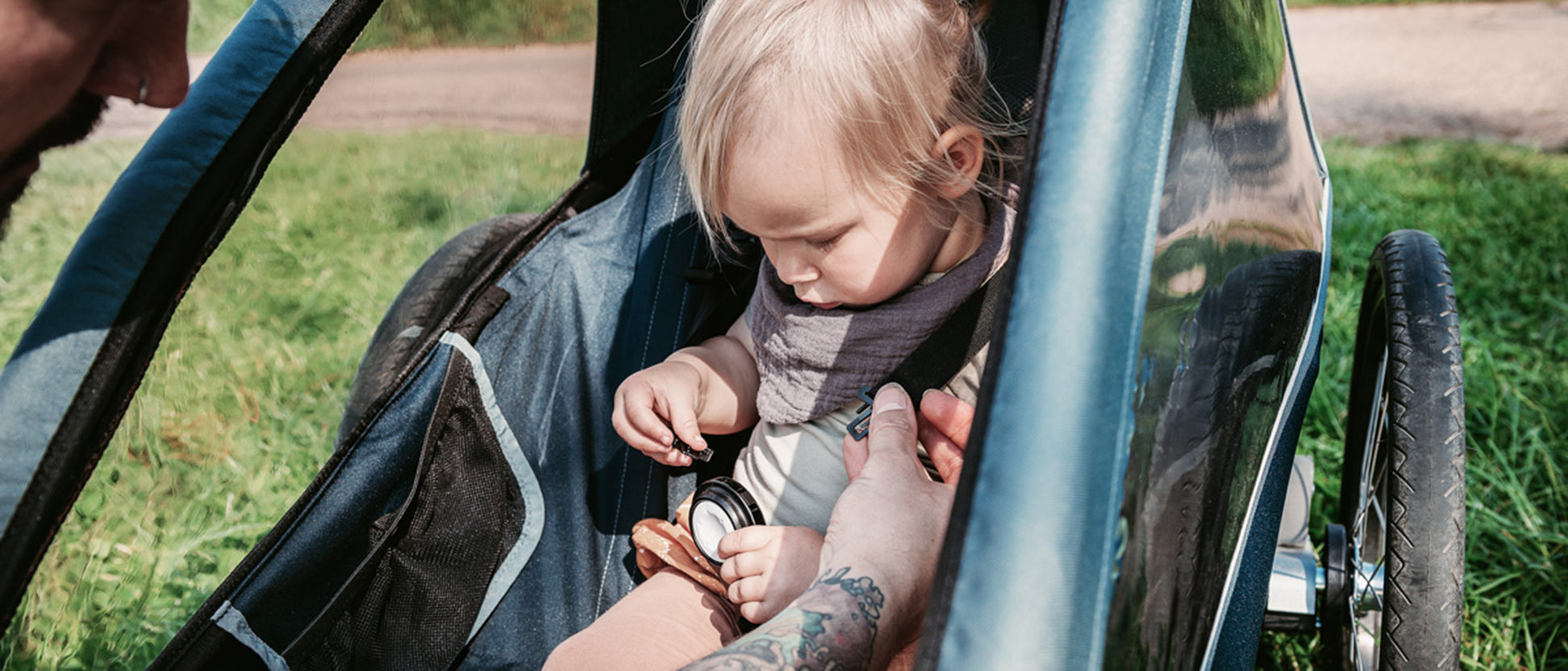 Dad secures his daughter with a 5-point harness inside a Croozer kid trailer; field path and meadow behind.